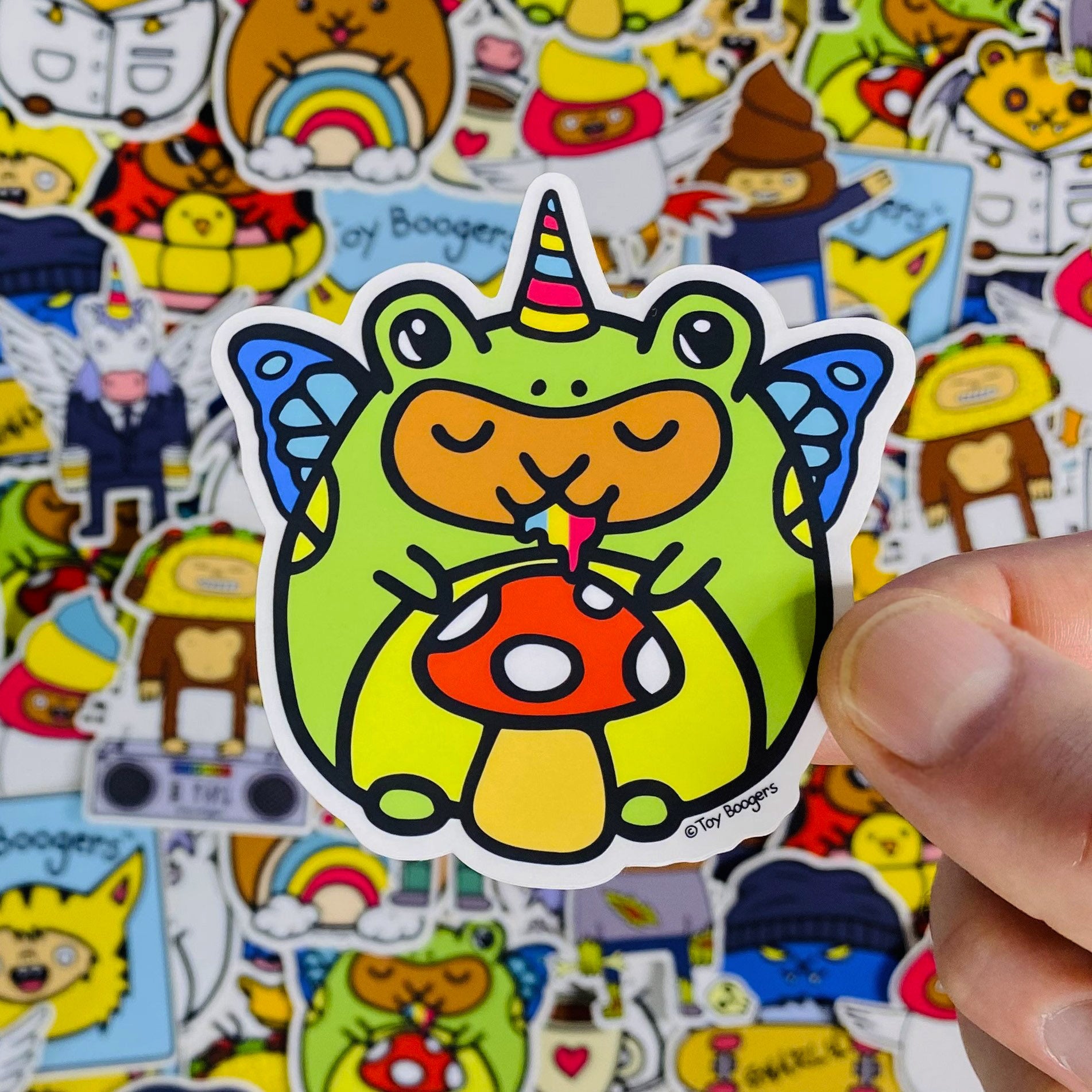 Whimsical sticker of a hamster wearing a frog costume with butterfly wings and a unicorn horn holding a mushroom.